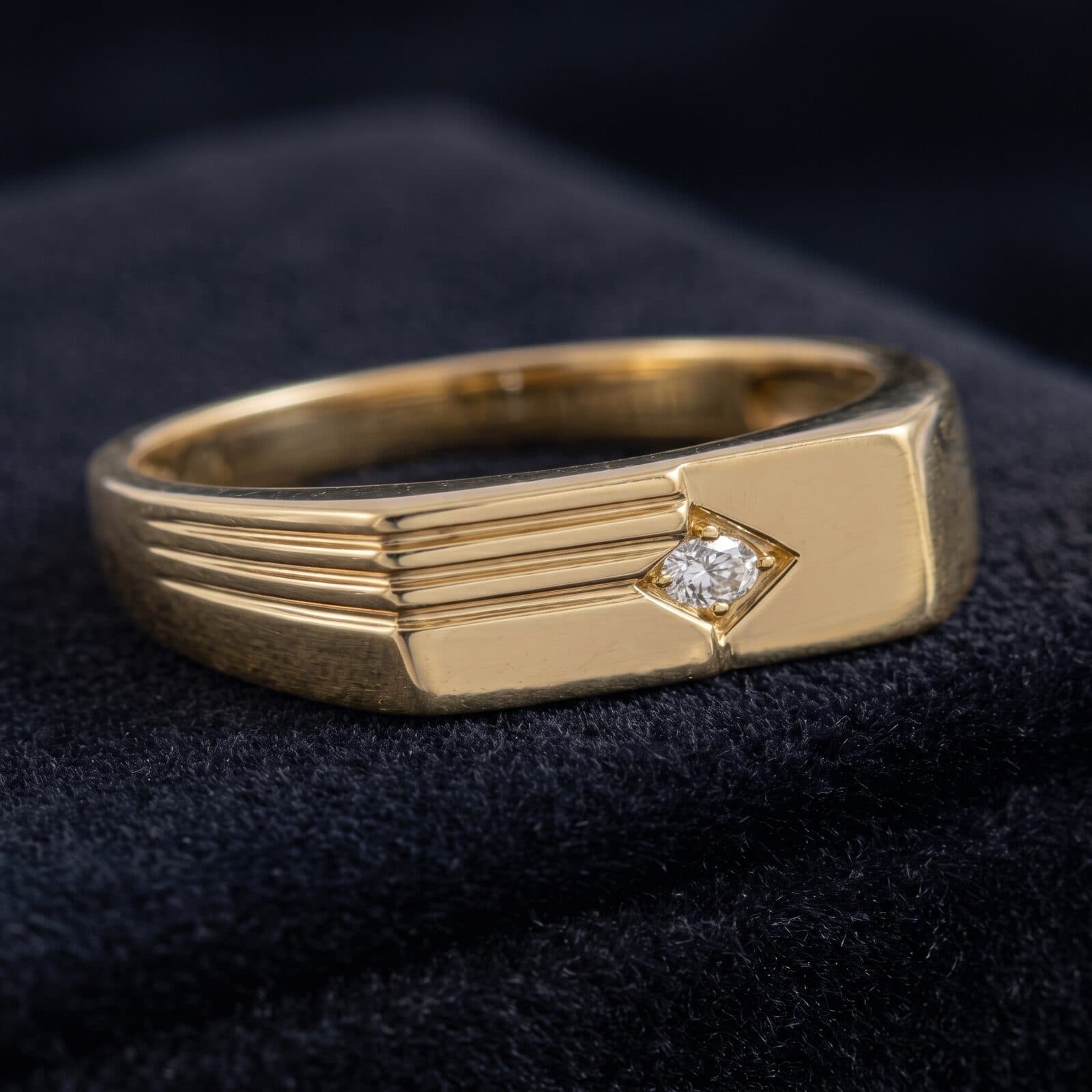 A men's gold signet ring with a ribbed top and a single diamond, on black velvet